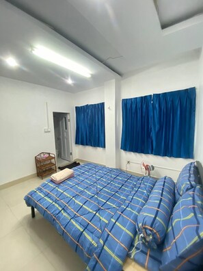 Ein Schlafzimmer mit einem Bett, einem Ventilator, einem Fenster mit blauen Vorhängen und einer Tür.