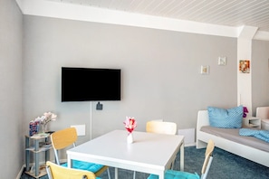 A modern room with a flat-screen TV, a white table with a vase of flowers, a sofa with patterned cushions, and a bookshelf with books and a lamp.