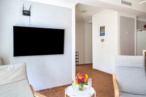 A modern living room with a flat-screen TV mounted on the wall, a round table with a vase of flowers, and two chairs.