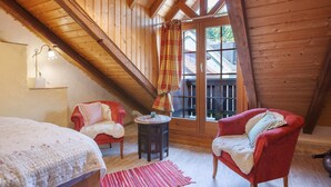 A cozy attic room with wooden beams, a red chair, a white chair, a small round table, and a window with a plaid curtain.
