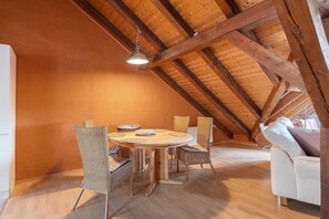 Un espace repas comprenant une table et des chaises en bois, un luminaire suspendu et un plafond en pente avec des poutres apparentes.