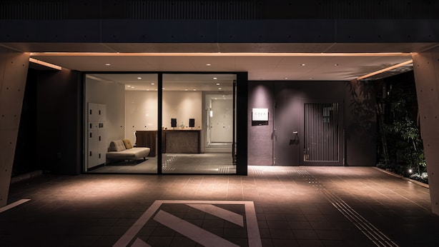 A modern hotel entrance with a reception desk, a sofa, and a glass door leading inside.