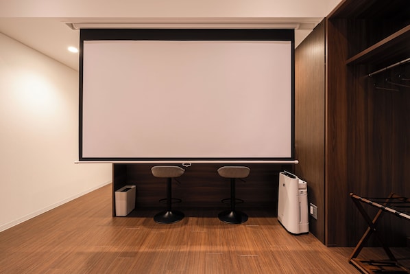 A room with a large projection screen, two bar stools, a trash bin, and a coat rack.