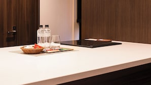 A modern kitchen with a white countertop, a stainless steel range hood, and wooden cabinetry. There are two bottles of water and a fruit bowl on the counter.