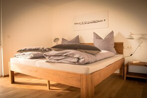 Een net opgemaakt bed met kussens en een deken, een nachtkastje met een lamp, en een aan de muur hangend kunstwerk.