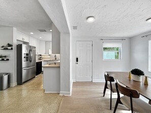 Una cocina moderna con un refrigerador de acero inoxidable, horno empotrado y lavavajillas. Hay una zona de comedor con mesa y sillas de madera.