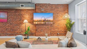 A modern living room with a brick wall, a sofa, a coffee table, a lamp, a chair, and a painting of a city skyline. - Charming 3-bedroom house in wonderful St. Louis with AC, WiFi (St. Louis)