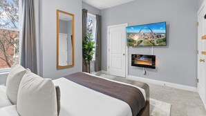 A hotel room with a bed, a TV mounted on the wall, a fireplace, and a window with curtains. - Charming 3-bedroom house in wonderful St. Louis with AC, WiFi (St. Louis)