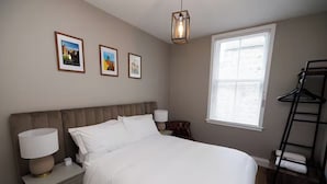 A bedroom with a bed, bedside lamps, a hanging light fixture, a window with blinds, and a wall-mounted rack. - Stylish 1-Bed Apartment in the Heritage Quarter (Dunfermline)