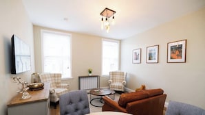 A living room with a sofa, armchairs, a coffee table, and a TV. There are framed pictures on the wall and a ceiling light fixture. - Stylish 1-Bed Apartment in the Heritage Quarter (Dunfermline)