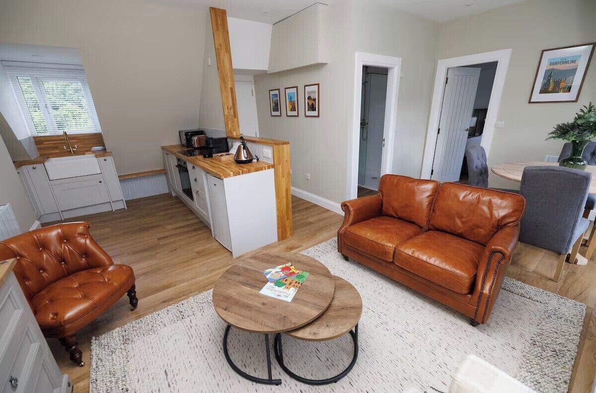 A modern living room with a brown leather sofa, a wooden coffee table, and a kitchen area with a wooden countertop and white cabinets.