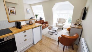 Cocina compacta con encimera de madera, armarios blancos y horno empotrado. Hay un comedor con una mesa redonda y dos sillas, además de una sala de estar con sofá y sillón.