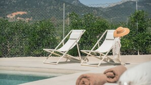 Two white lounge chairs by a pool with a mountain backdrop, a fence, and a rolled towel on the ground. - Can Cama Rotja, pool, barbecue & incredible views (Inca)