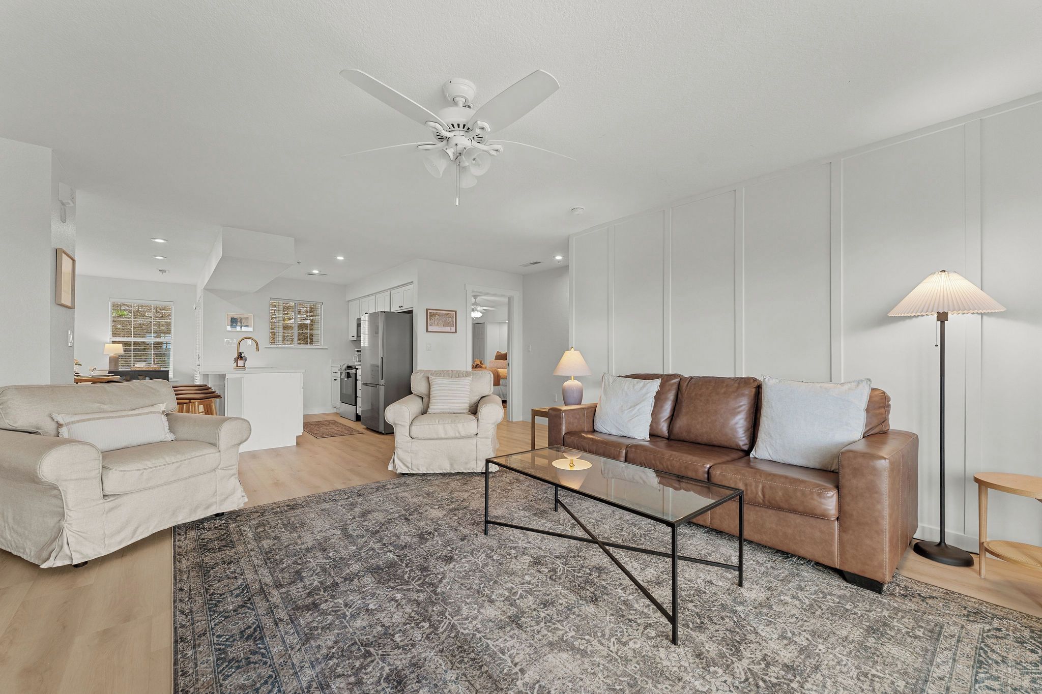 A spacious living room with a ceiling fan, a brown leather sofa, a white armchair, and a glass coffee table.