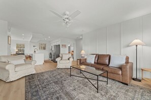 A spacious living room with a ceiling fan, a brown leather sofa, a white armchair, and a glass coffee table. - Gated Lake View Condo W/ pool, private boat ramp, putt putt, views of lake (Kimberling City)