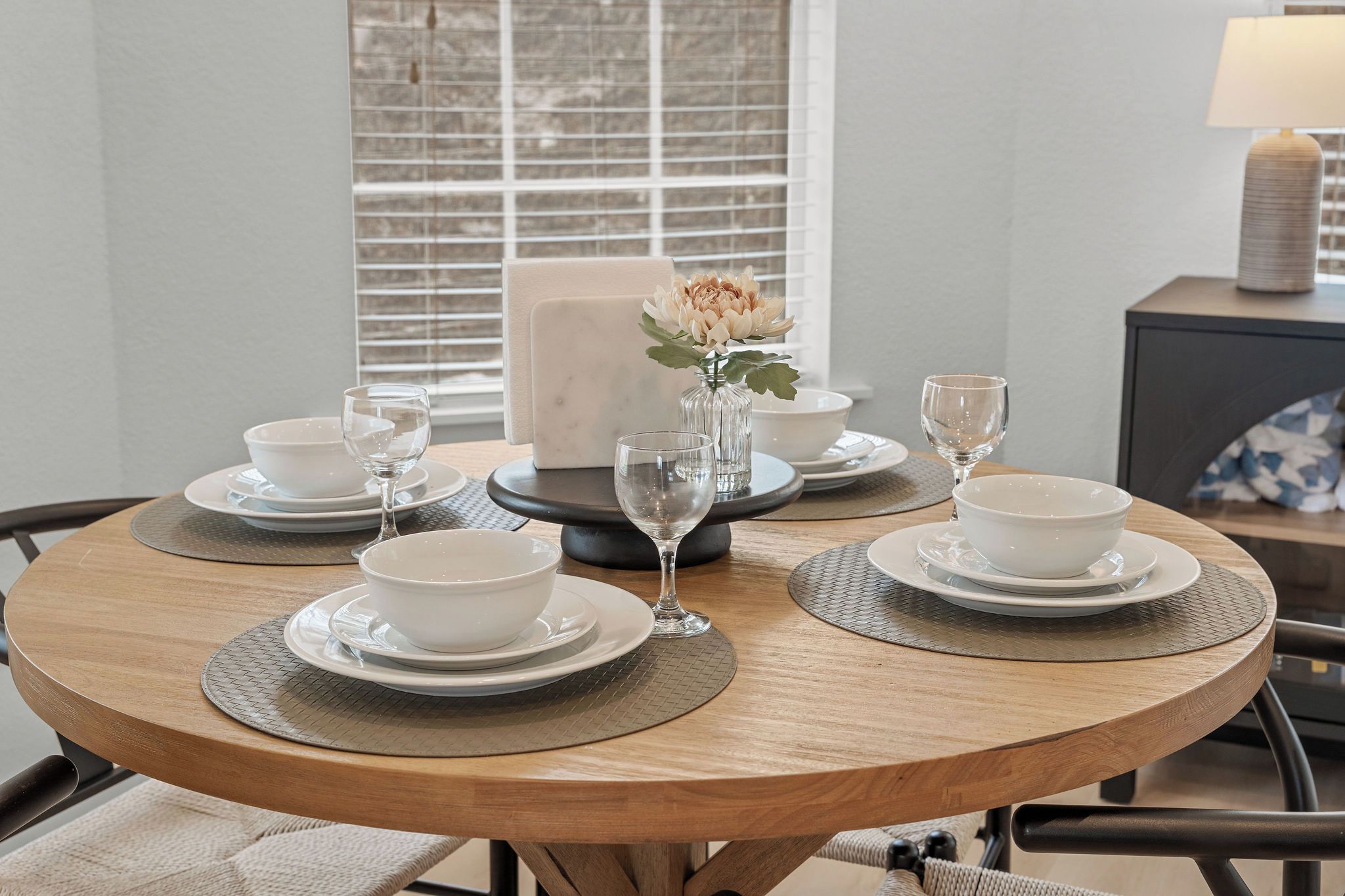 A round dining table with white plates, bowls, and wine glasses, set with a vase of flowers.