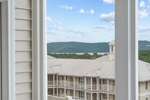 A view through a window of a building with a balcony overlooking a body of water and a forested hill. - Gated Lake View Condo W/ pool, private boat ramp, putt putt, views of lake (Kimberling City)