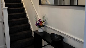 Un escalier avec des marches recouvertes d’une moquette sombre mène à un palier. Une console noire, sur laquelle est posé un vase de fleurs, se trouve sur le palier. Un grand miroir est accroché au mur, au-dessus de la console.