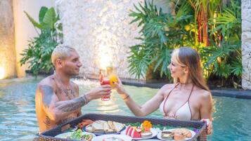 Two people toasting with drinks in a poolside setting with a wicker tray of food and a stone wall in the background.