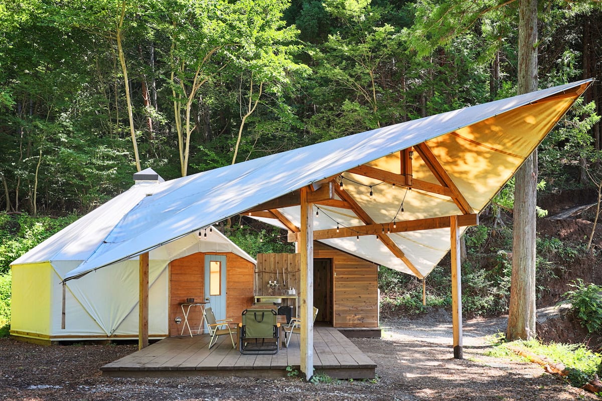 A rustic cabin with a large tent and a wooden deck, surrounded by dense forest.