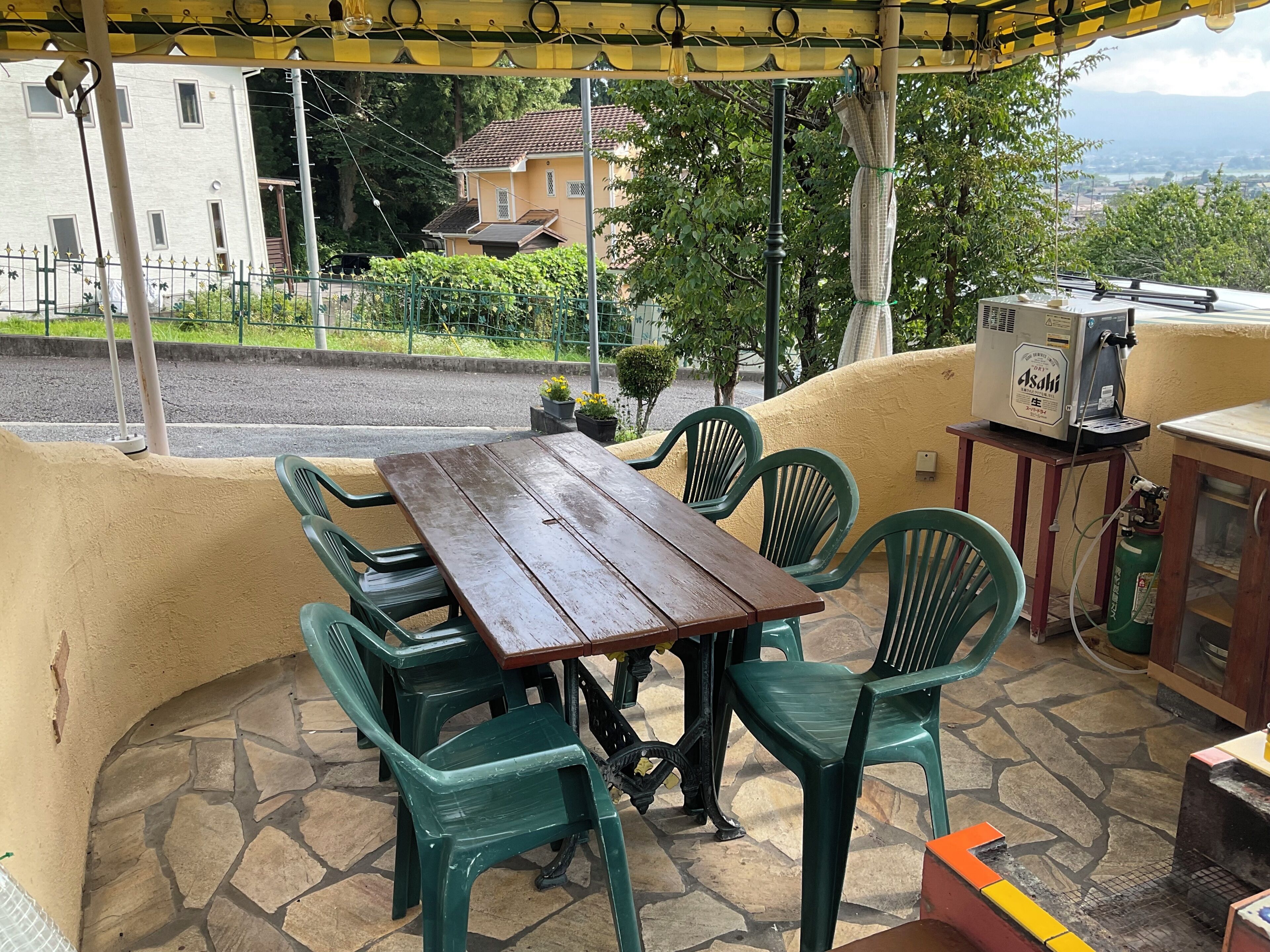 A covered outdoor seating area with a wooden table and green plastic chairs, a gas stove, and a water dispenser.
