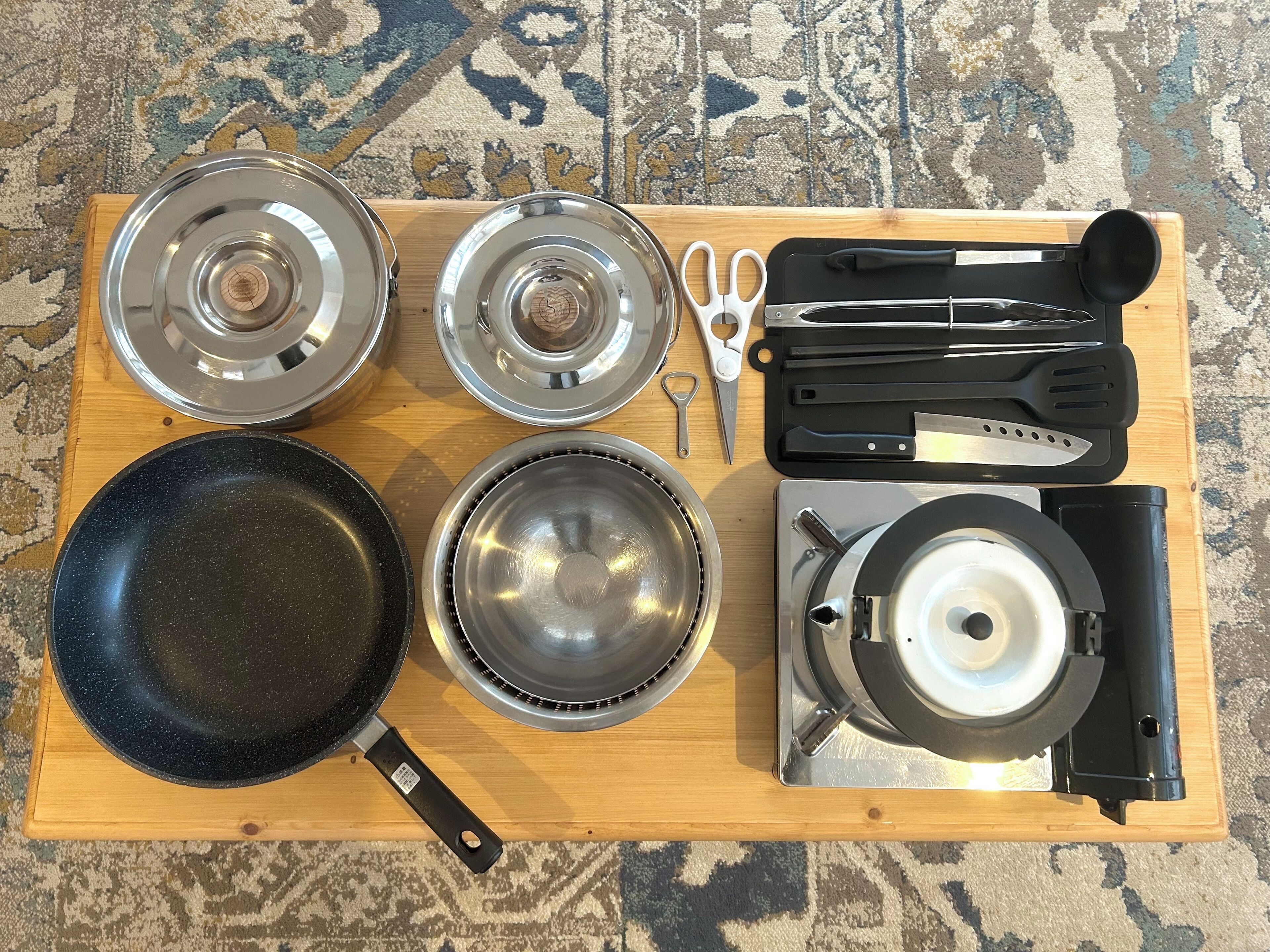 A wooden cutting board with various kitchen utensils and cookware, including a frying pan, lids, a pair of scissors, and a grater.