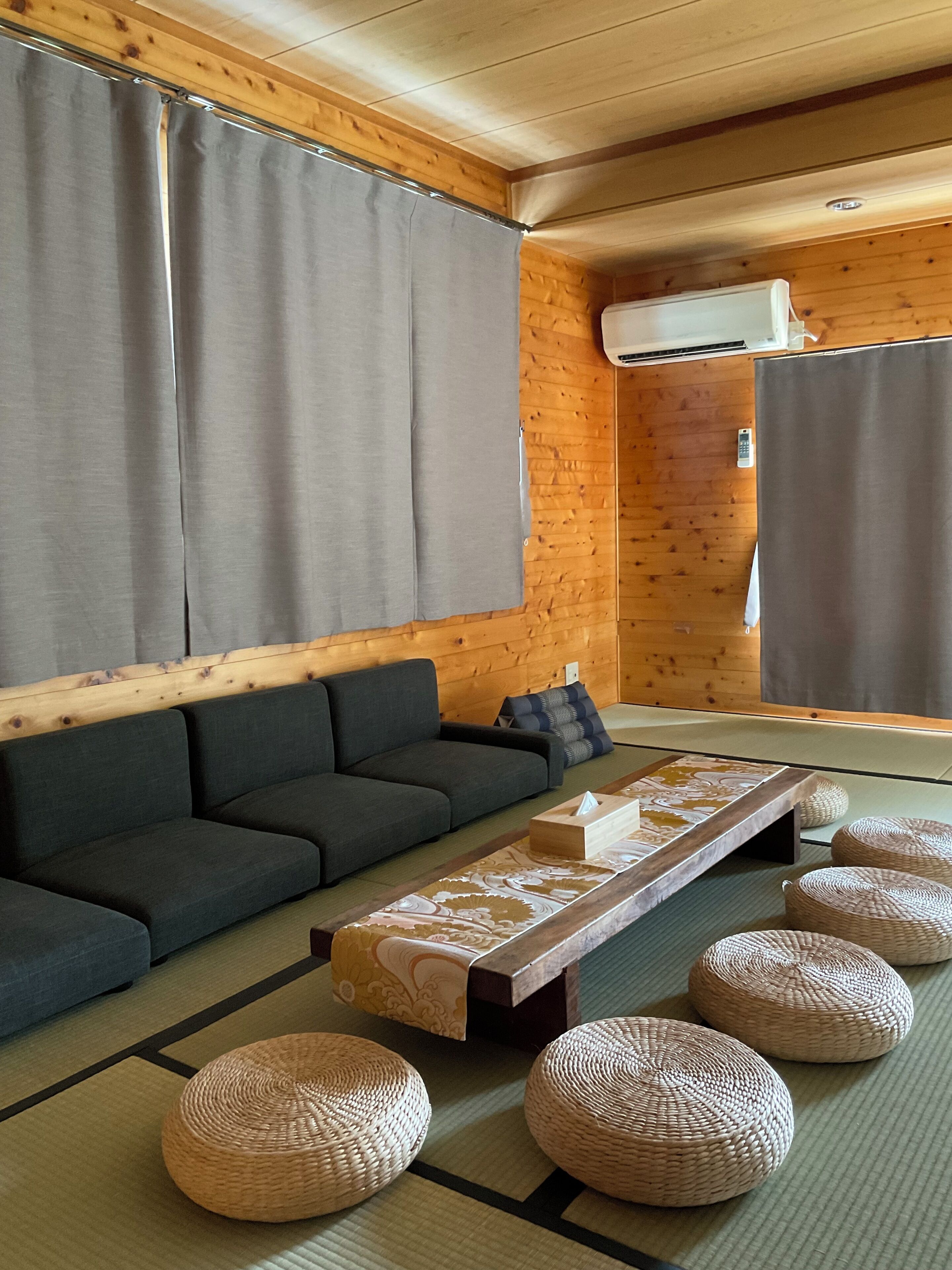 A room with a dark sofa, a low table with a patterned runner, and woven poufs on tatami flooring.