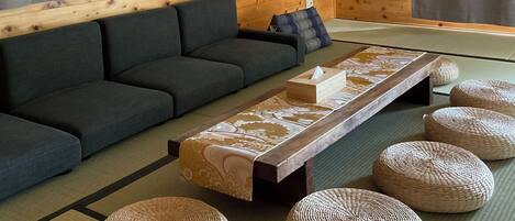 A room with a dark sofa, a low table with a patterned runner, and woven poufs on tatami flooring.