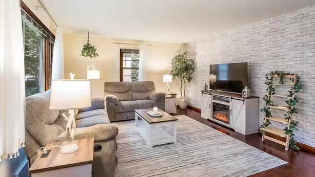 Una sala de estar moderna con sofá, mesa de centro y televisor, que presenta una pared de ladrillo y ventanas amplias.