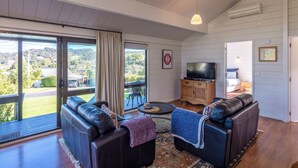 A spacious living room with a large sliding glass door leading to a balcony, a flat-screen TV, and a cozy seating area with leather sofas. - Matai Cottage in Oneroa with Sea Views (Waiheke Island)