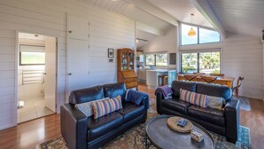 A spacious living area with two black leather sofas, a coffee table, and a dining table with chairs. There are large windows providing natural light and a view of the outdoors. - Matai Cottage in Oneroa with Sea Views (Waiheke Island)