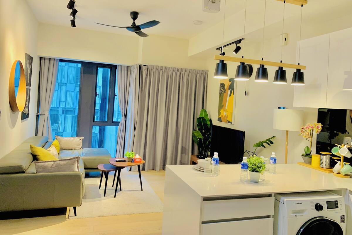 A modern living room with a sofa, a coffee table, and a dining area. There is a ceiling fan, a TV, and a washing machine in the kitchen area.