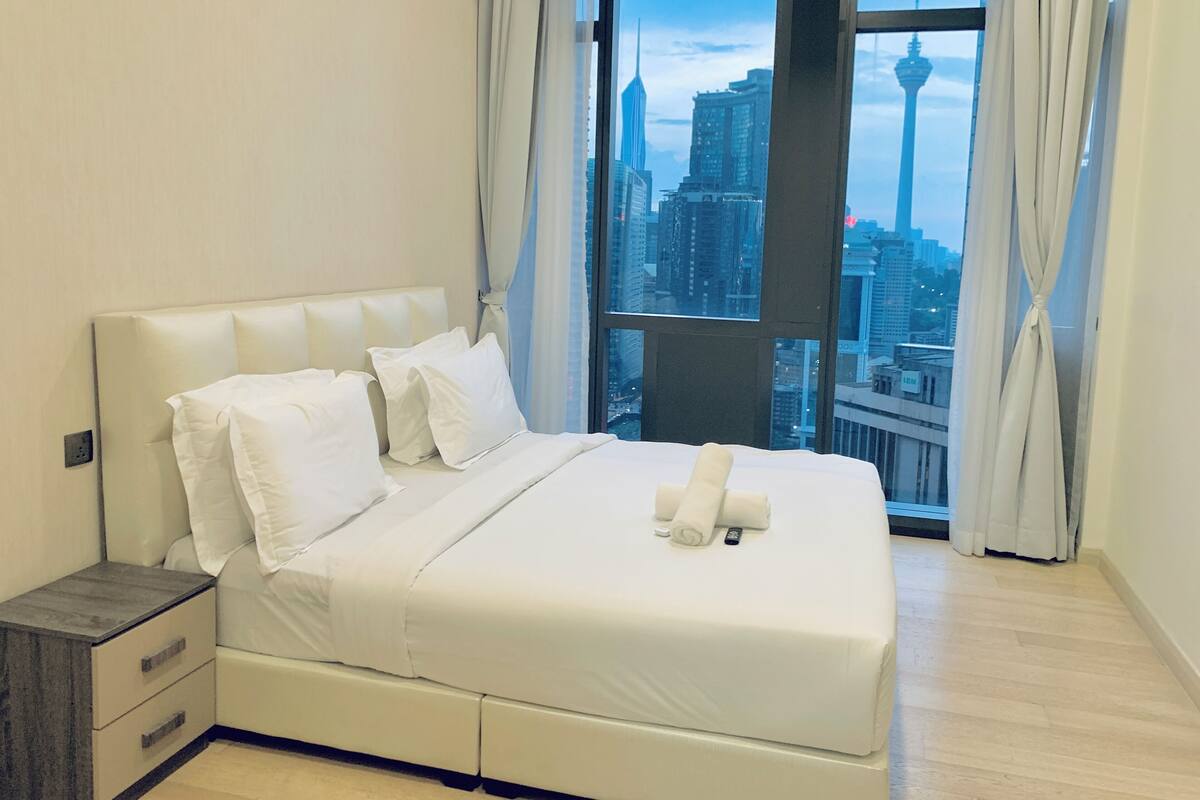 A modern hotel room with a large bed, a nightstand, and a view of a cityscape through large windows.