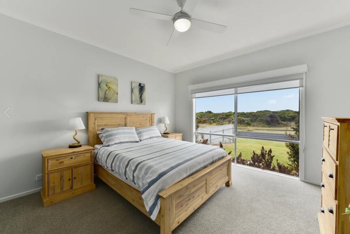 Quarto com uma grande janela, cama de madeira, mesa de cabeceira e cômoda.
