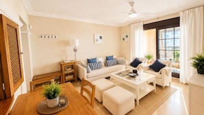 A living room with a sofa, coffee table, and a dining table with chairs. There is a ceiling fan, a window with curtains, and a door with a wooden lattice. - Los Pelícanos - Ref.412 (Punta del Moral)