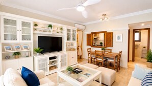 A living room with a TV, white furniture, a dining area, and a kitchen visible through an open door. - Los Pelícanos - Ref.412 (Punta del Moral)