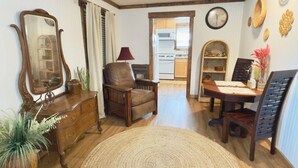 A living room with a wooden floor, a round woven rug, a wooden dining table with chairs, a wooden cabinet, a mirror, and a kitchen visible through an open doorway.
