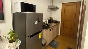 A compact kitchen with a stainless steel refrigerator, a microwave, and a sink. There is a potted plant on a small table, and a wooden door in the background.