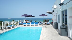 Un espace piscine avec des chaises longues bleues et des parasols, une vue dégagée sur l’océan et un panneau indiquant les heures d’ouverture de la piscine.