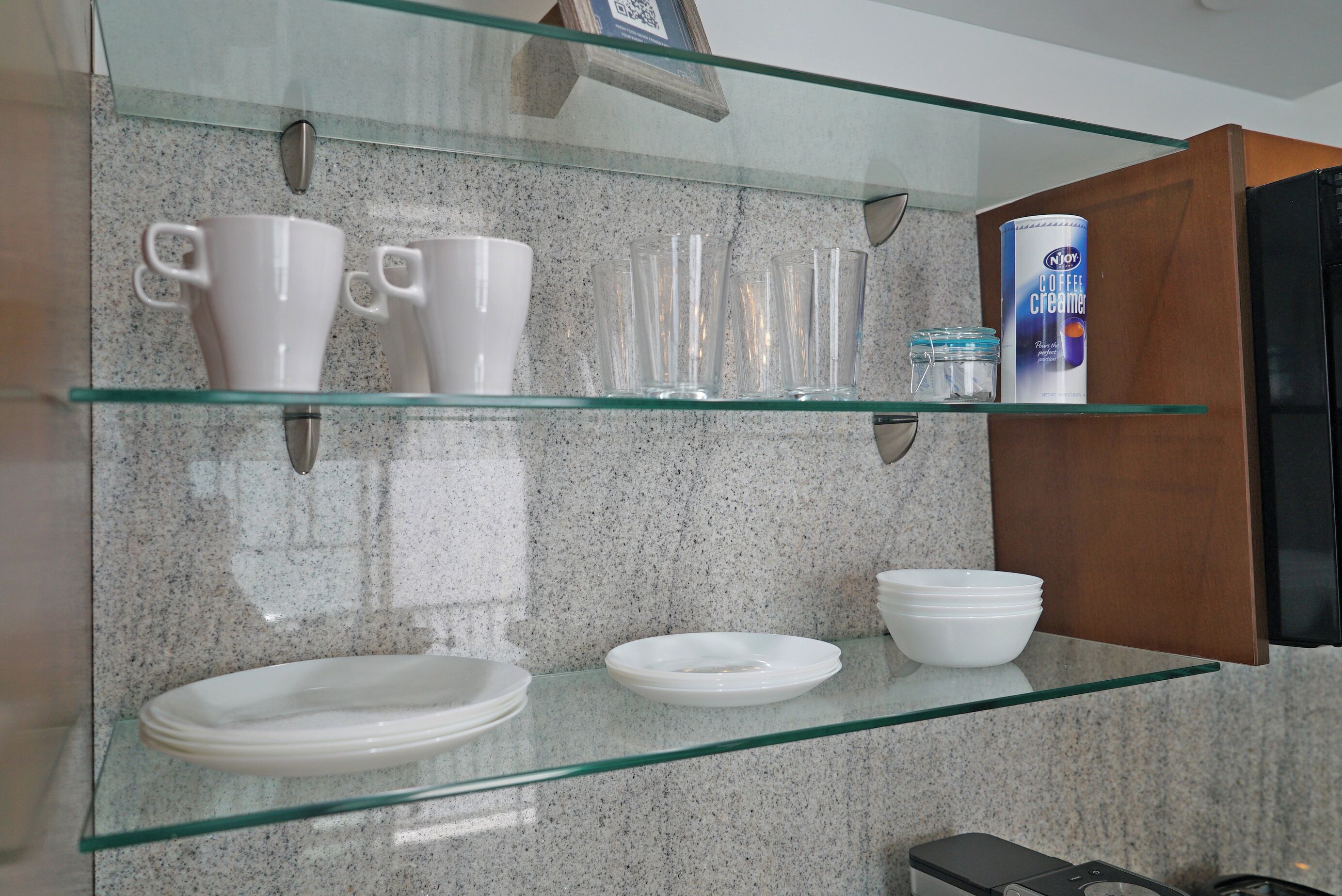 Una cocina con repisas de vidrio que sostienen tazas, vasos y platos blancos, además de una lata de crema de café.