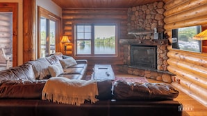 A log cabin interior with a leather sofa, stone fireplace, and a large window overlooking a lake.