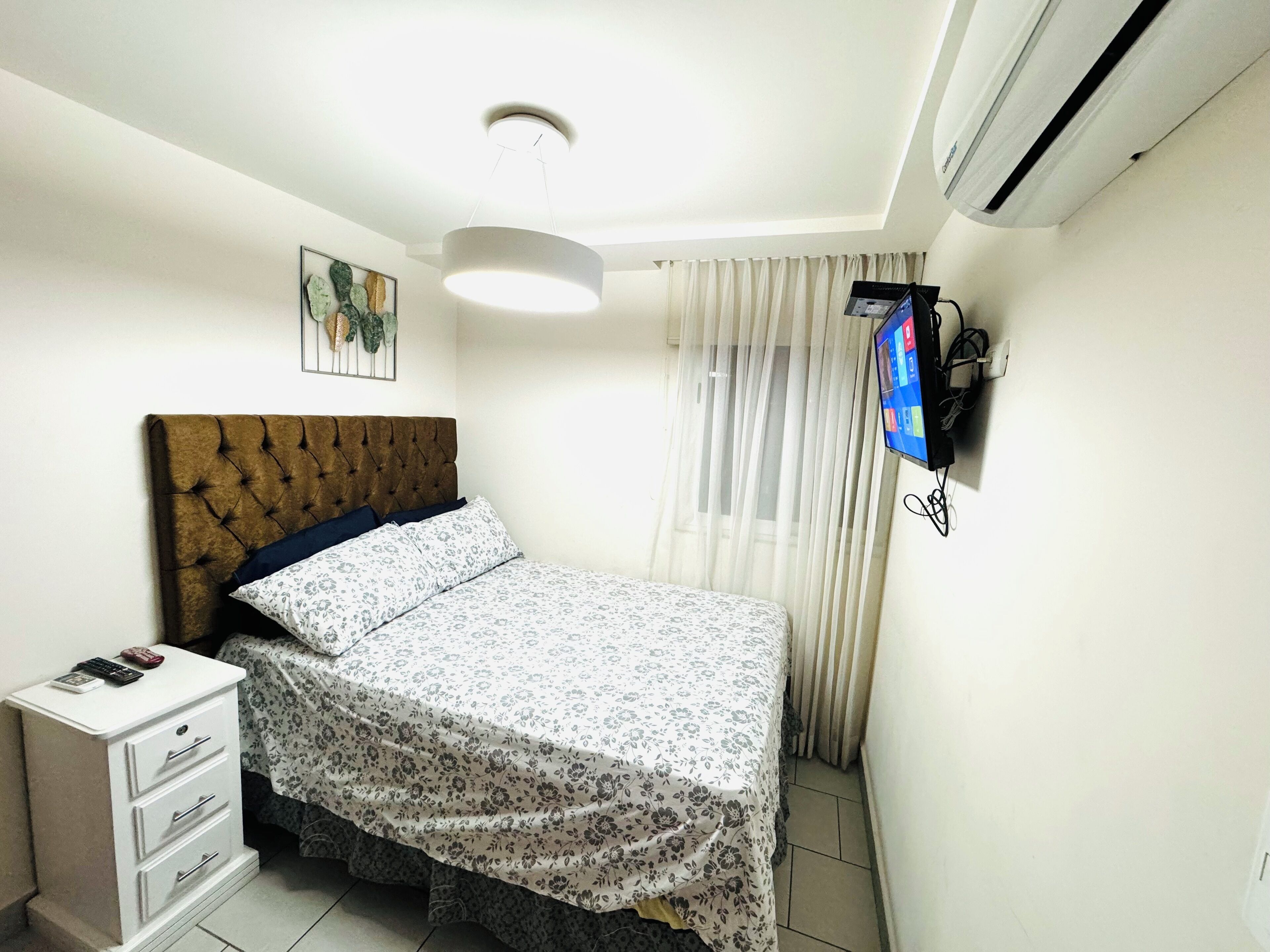 Ein Schlafzimmer mit einer geblümten Polsterkopfteile, einem Bett mit floralem Bettzeug, einem Nachttisch mit Schubladen, einem Wand-Fernseher und einer Deckenleuchte.