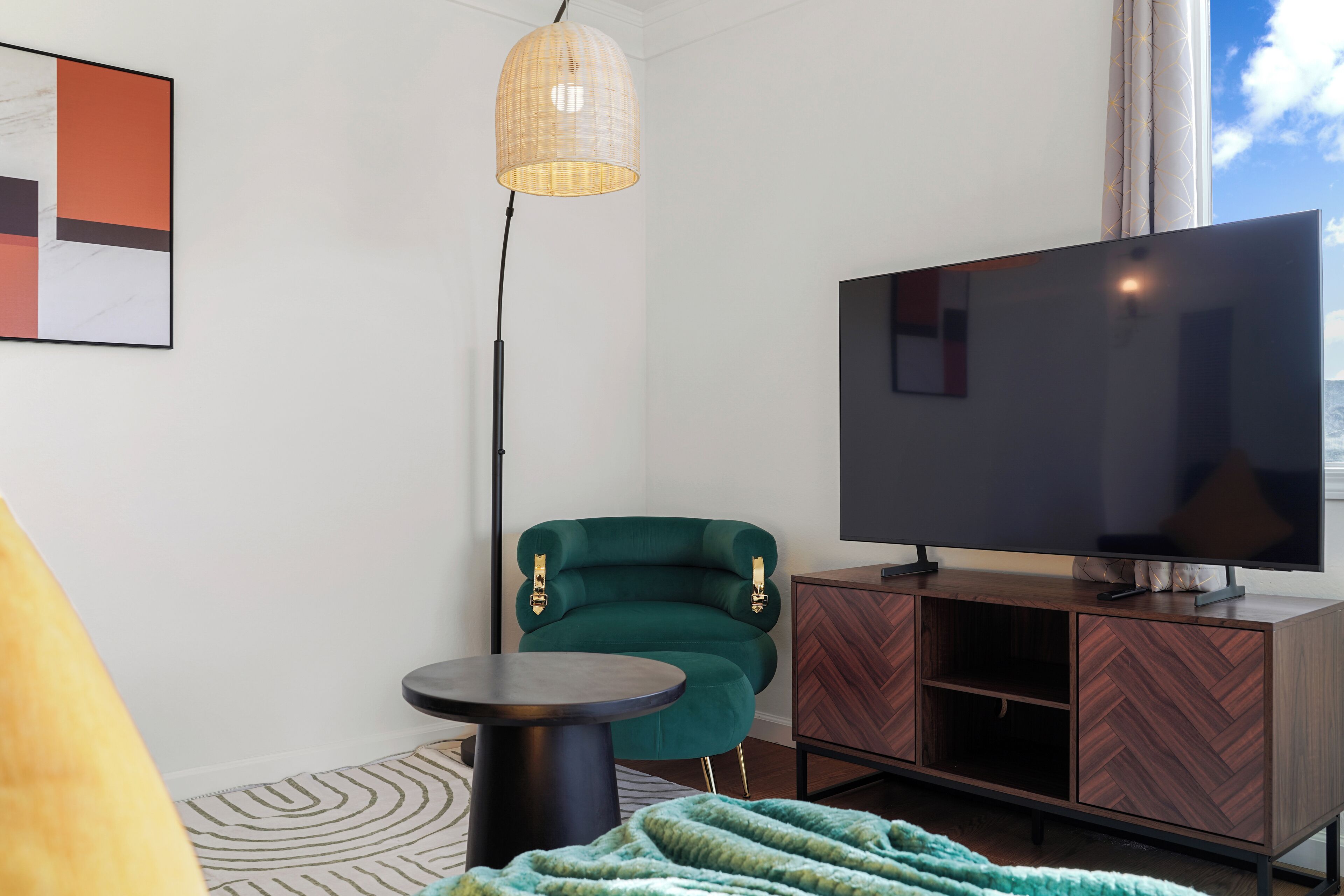 A modern living room with a large flat-screen TV, a green armchair, a small round table, and a herringbone-patterned wooden cabinet.
