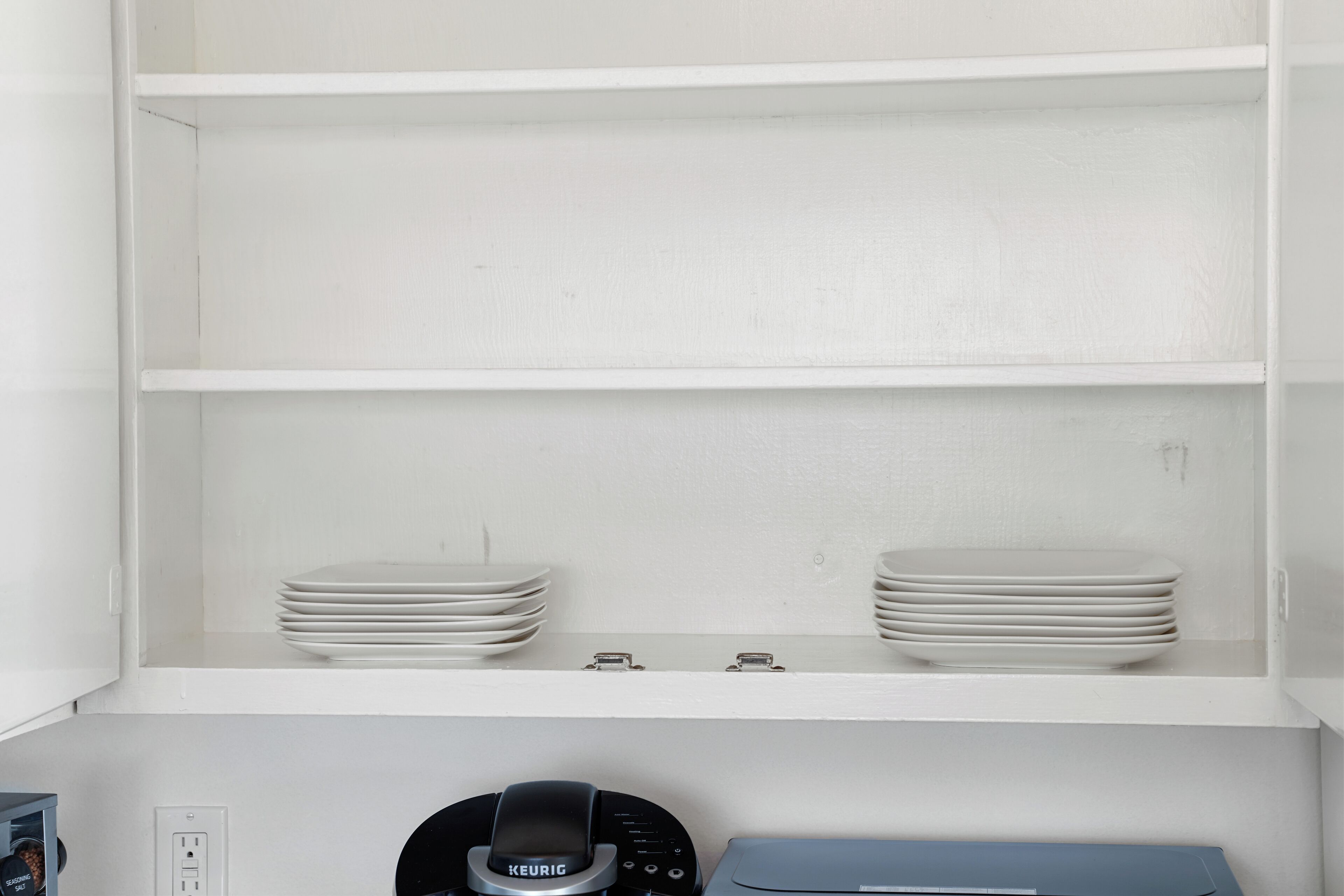 Ein weißes Regal mit gestapelten weißen Tellern und einer schwarzen Kaffeemaschine mit dem Aufdruck KEURIG.