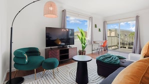 A modern living room with a flat-screen TV, a green armchair, a round coffee table, and a large window offering a view of the outdoors. - Charming 2BR Coastal Getaway in Pacifica (Pacifica)