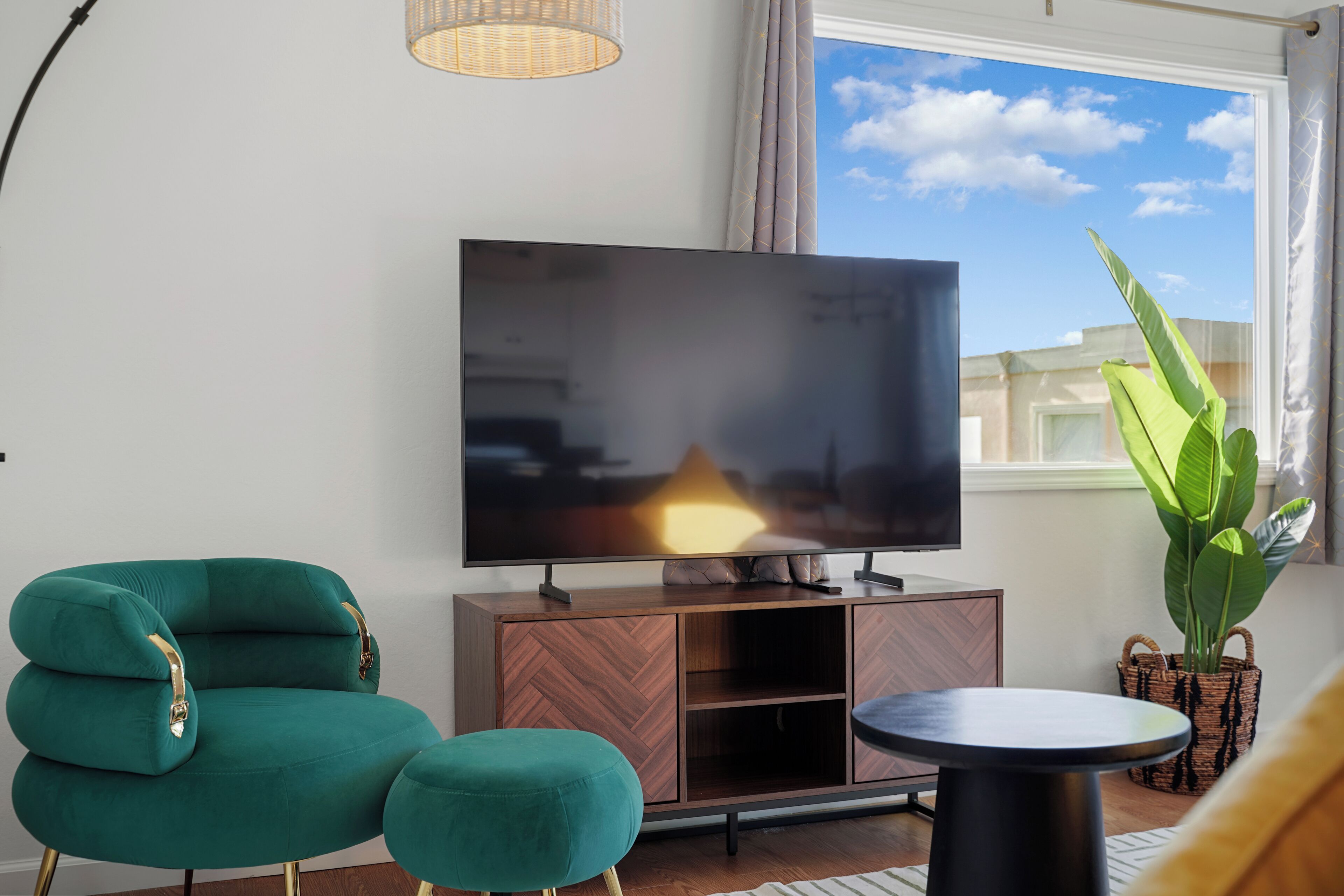 A modern living room with a flat-screen TV on a wooden cabinet, teal upholstered armchair, ottoman, and a small round table.