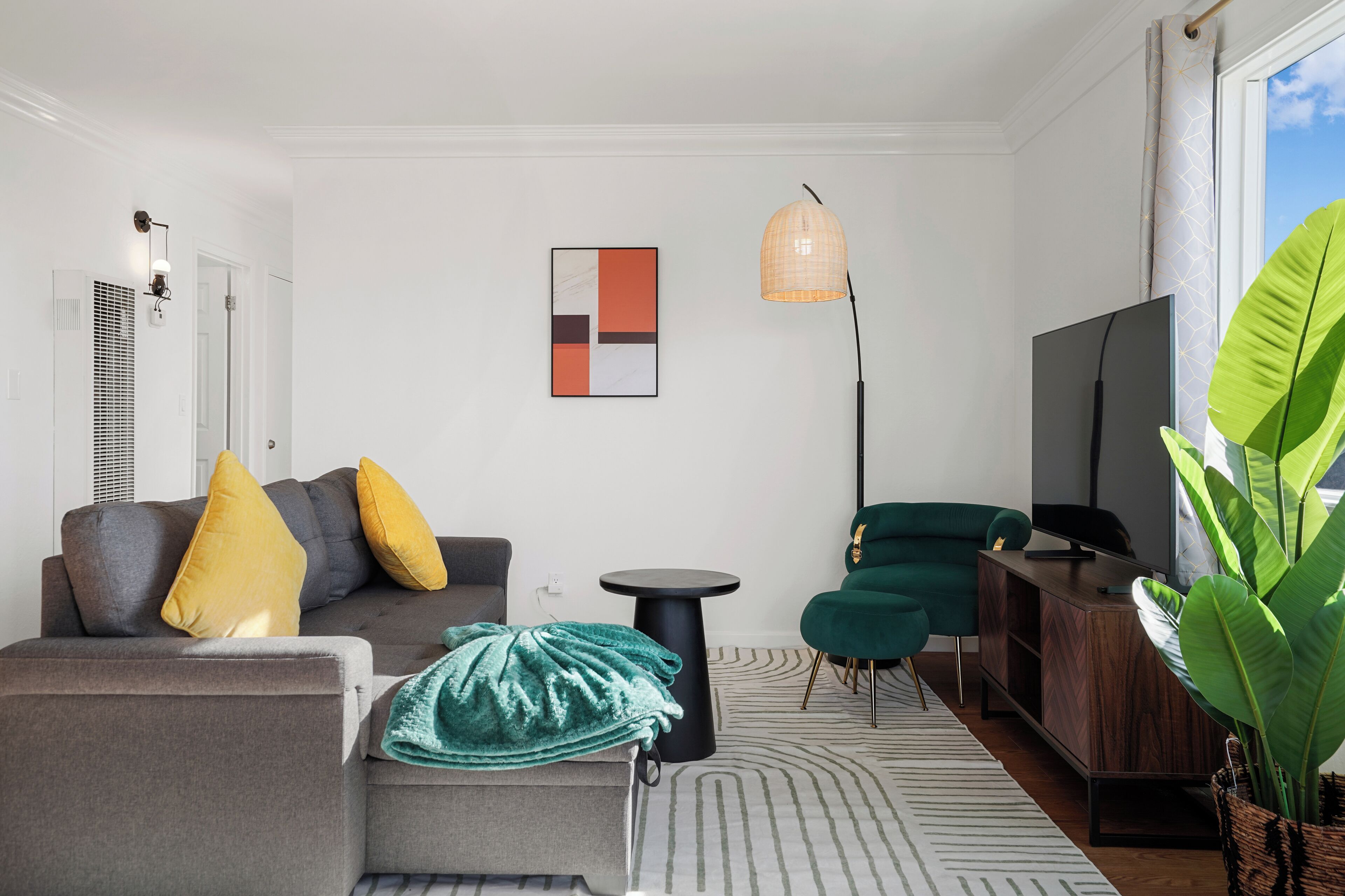 Un salon moderne avec un canapé gris, des coussins jaunes et verts, une petite table d’appoint noire, un fauteuil vert, un meuble en bois et une grande fenêtre donnant sur un beau paysage.
