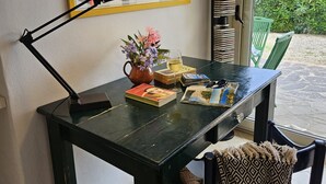 Un espacio de trabajo con un escritorio, una lámpara, un jarrón con flores, una copa de vino y un cartel enmarcado en la pared.