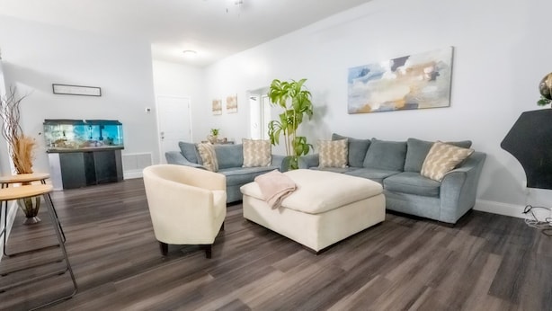 A modern living room with a grey sofa, a cream-colored ottoman, and a white armchair. There is a large window with a view of the outdoors, a ceiling fan, and a flat-screen TV mounted on the wall.