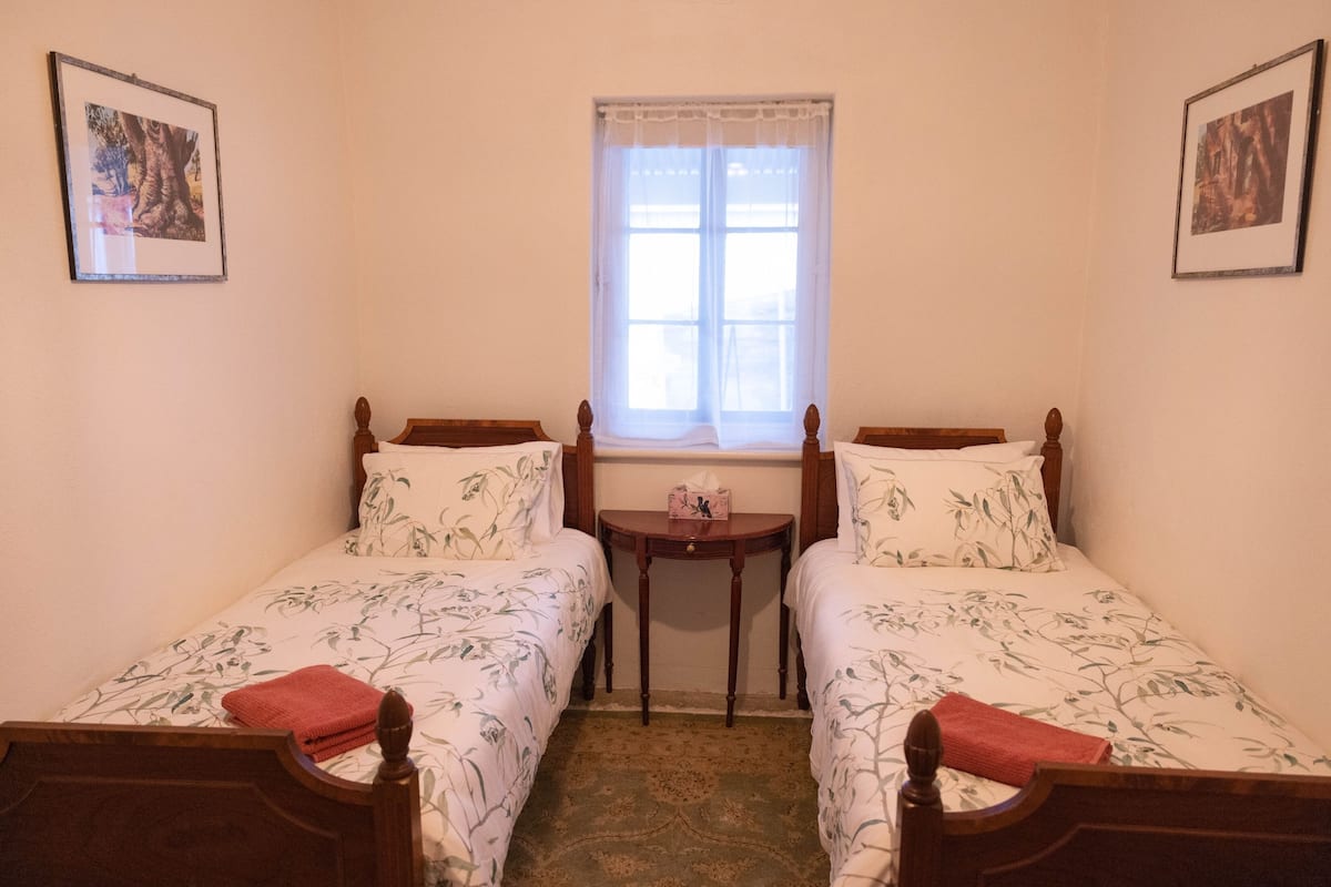 Una habitación pequeña y acogedora con dos camas individuales, una mesita y una ventana con cortina translúcida.